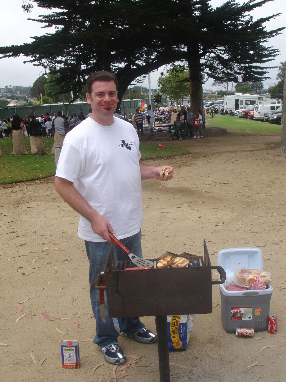 A man grilling hot dogs and buns at an outdoor picnic.