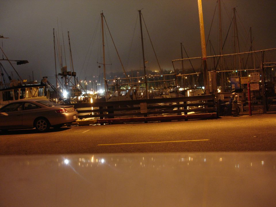 Silver sedan parked at a dock at night, with sailboats in the background.