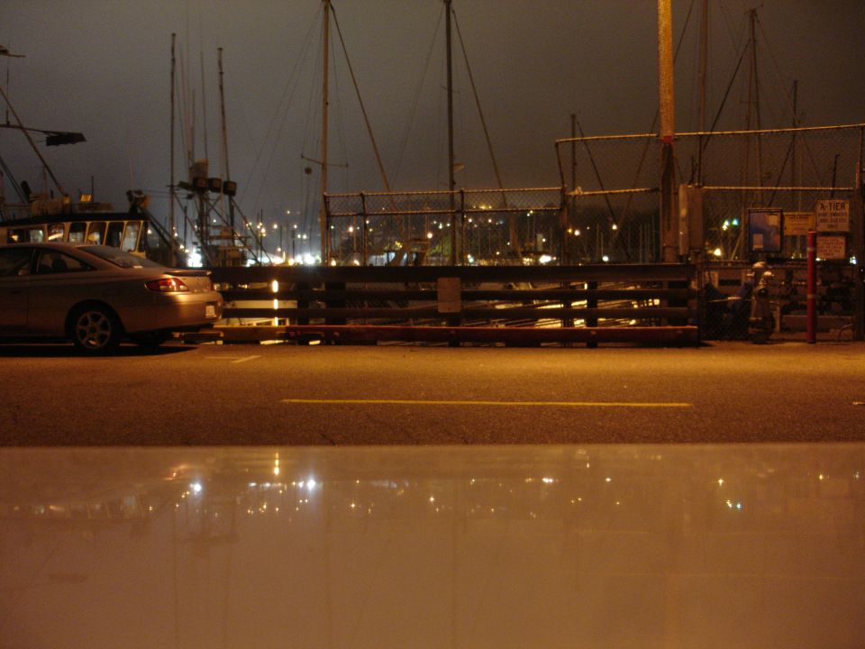 Silver sedan parked at night by a harbor with reflections in the wet ground.