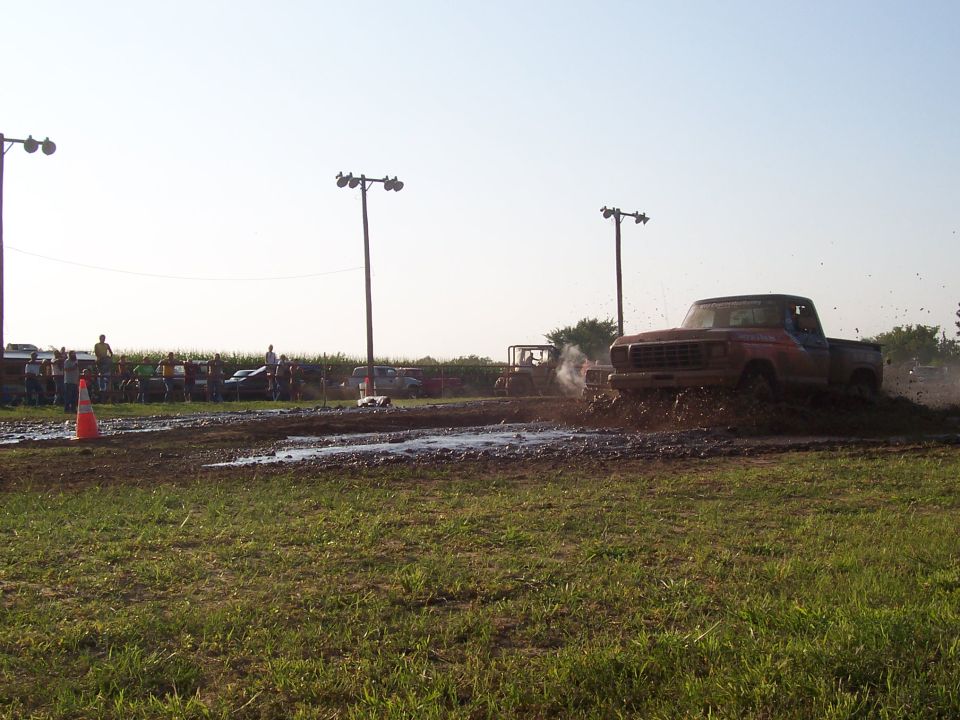 Red pickup truck stuck in mud pit with spectators in background.