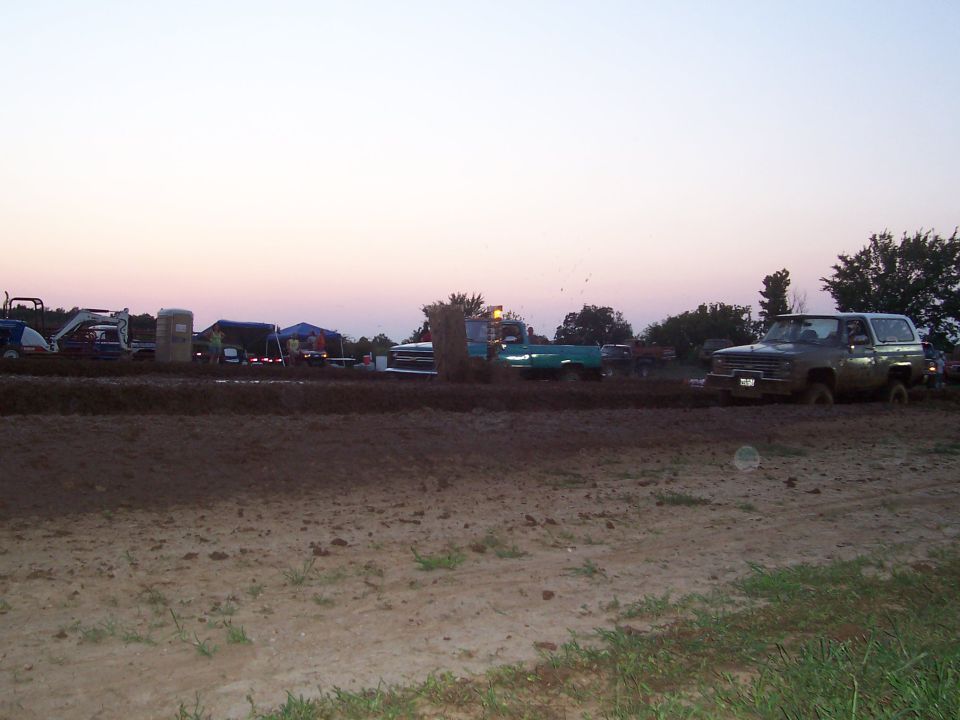 Dark SUV driving through mud pit with tents and other vehicles in background.