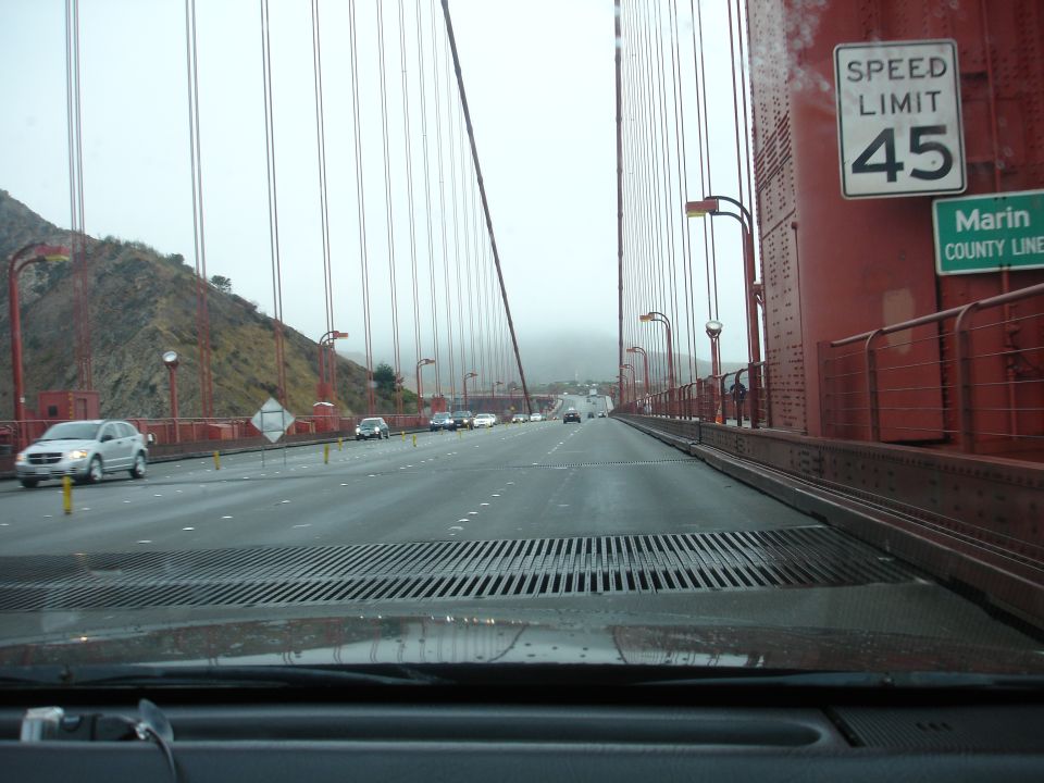View from inside a car driving on the Golden Gate Bridge, showing fog, red cables, and speed limit signs.