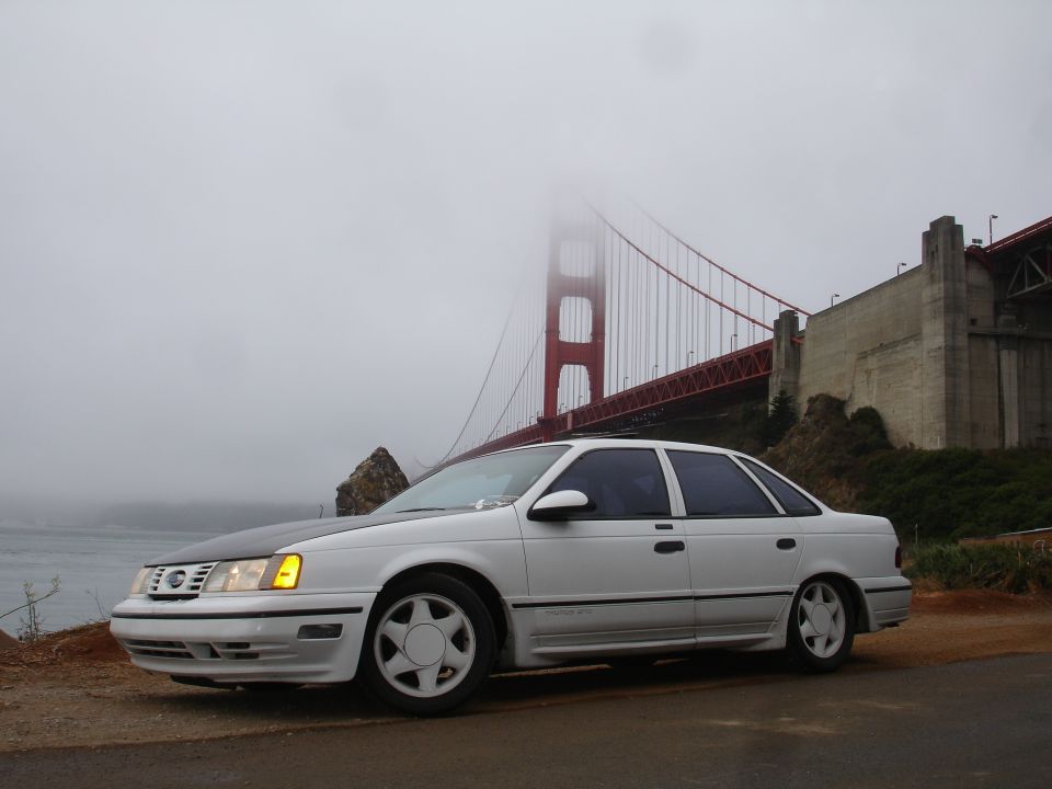 White 1990 Ford Taurus SHO sedan with black hood parked near Golden Gate Bridge on a foggy day.