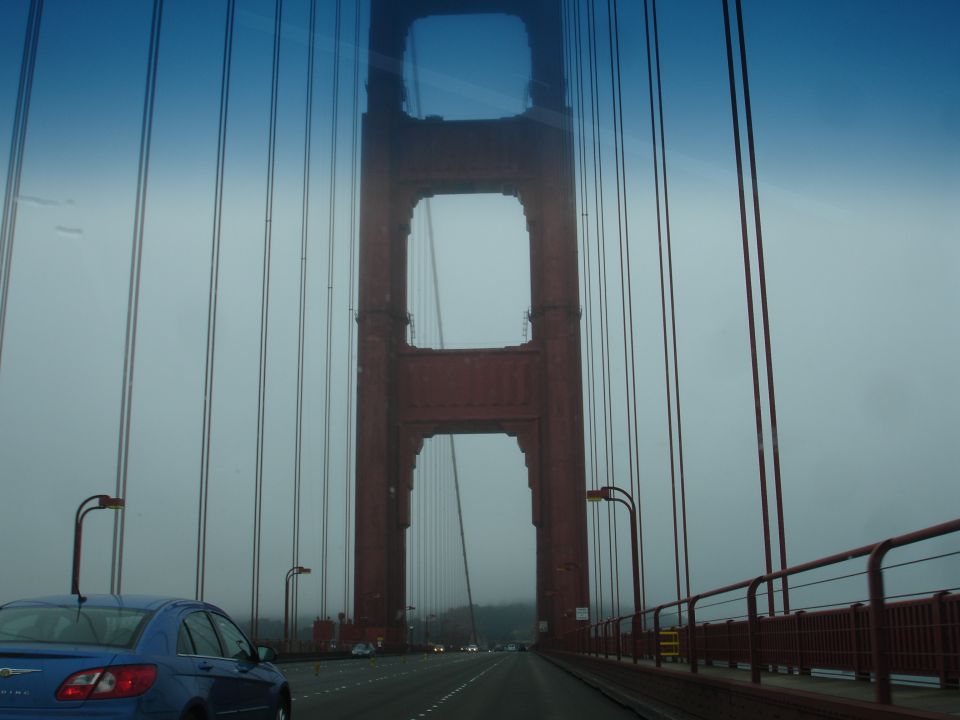 Blue sedan driving on Golden Gate Bridge with fog and red towers.