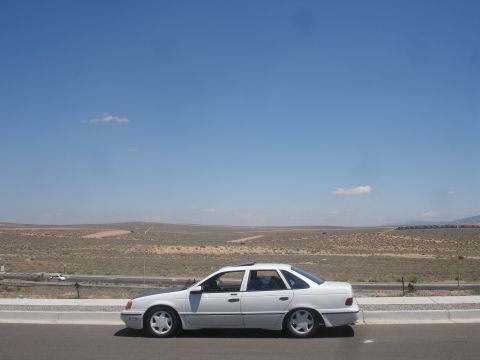 1990 Ford Taurus - A white sedan is parked on the side of the road with a vast desert landscape under a clear blue sky.