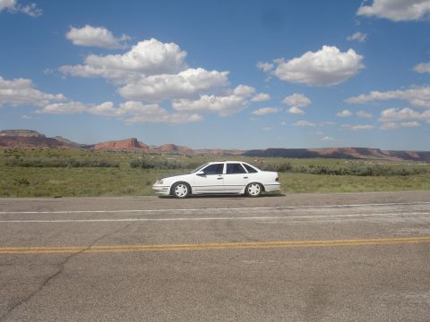 1990 Ford Taurus - A white 1990 Ford Taurus sedan is parked on the shoulder of a highway, with a scenic desert landscape under a cloudy blue sky.
