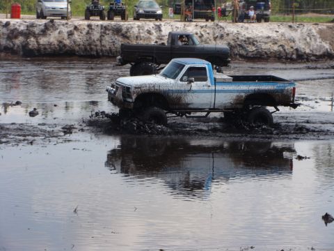 1982 Ford F-100 - A white and blue pickup truck is driving through a muddy track, kicking up a lot of mud. Another dark colored pickup truck is visible behin…