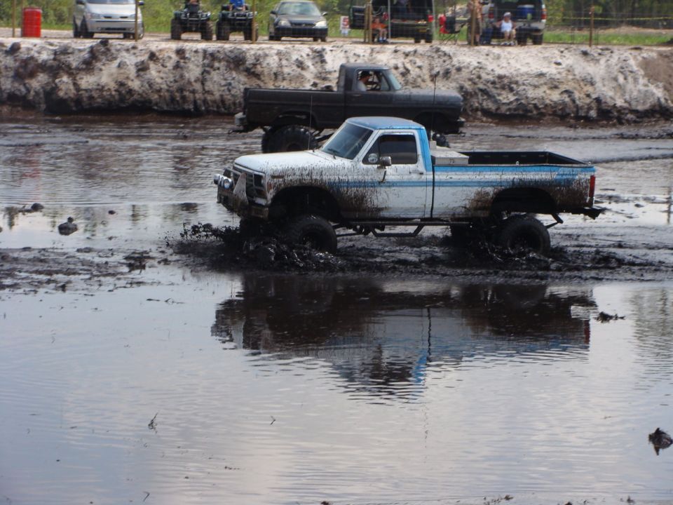 White and blue pickup truck driving through mud, with another truck behind it.