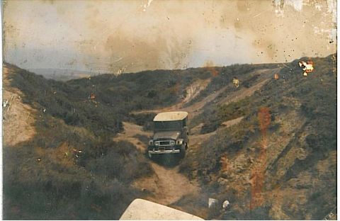 1991 Ford Bronco - A dark-colored, soft-top SUV drives down a dirt path between overgrown hills. The vehicle is seen from a front 3/4 angle, heading away from…