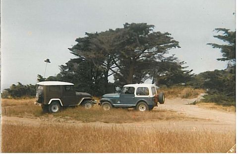 1991 Ford Bronco - Two vintage Jeep-style vehicles are parked on a dirt road next to tall grass and trees. A dark colored Jeep is on the left, and a blue and…