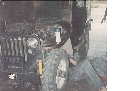 1991 Ford Bronco - A person in jeans is working underneath the engine of a dark-colored Jeep, which has its hood open. The Jeep has large tires and a visible…