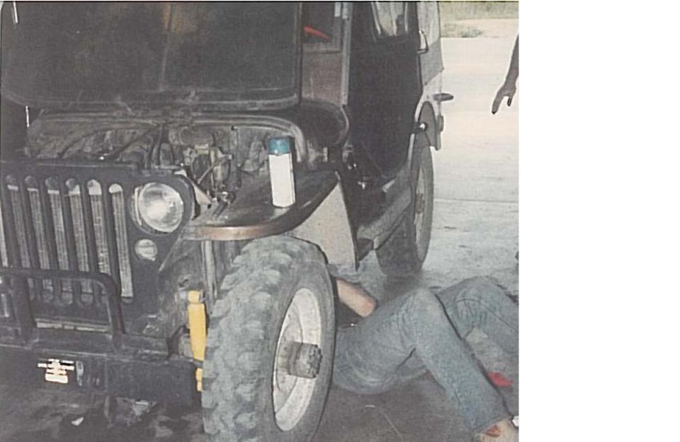 Person working on engine of a dark Jeep with hood open, large tires visible.