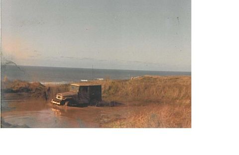 1991 Ford Bronco - Faded, grainy film photo of a dark 4x4 wagon with a white roof and whip antenna sitting in a muddy water-filled rut on a coastal dune trail…