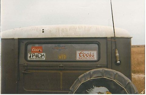 1991 Ford Bronco - Faded film photo of the rear of an olive-drab vintage panel truck or wagon with a white padded roof, its rear window covered in Coors Racin…