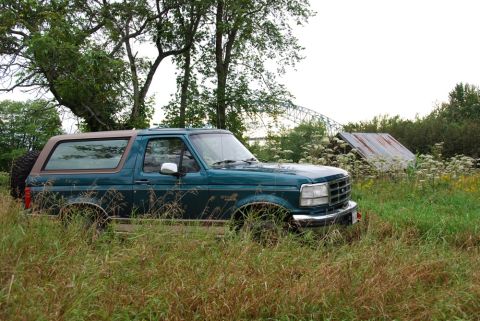 1996 Ford Bronco - Photograph of a 1990s teal Ford Bronco sitting in an overgrown grassy field, not a technical document.