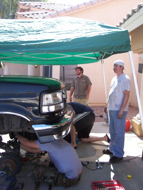 1996 Ford Bronco - Three men work on the front end of a black Ford Bronco, which is lifted by a blue jack. Tools and parts are scattered on the concrete groun…