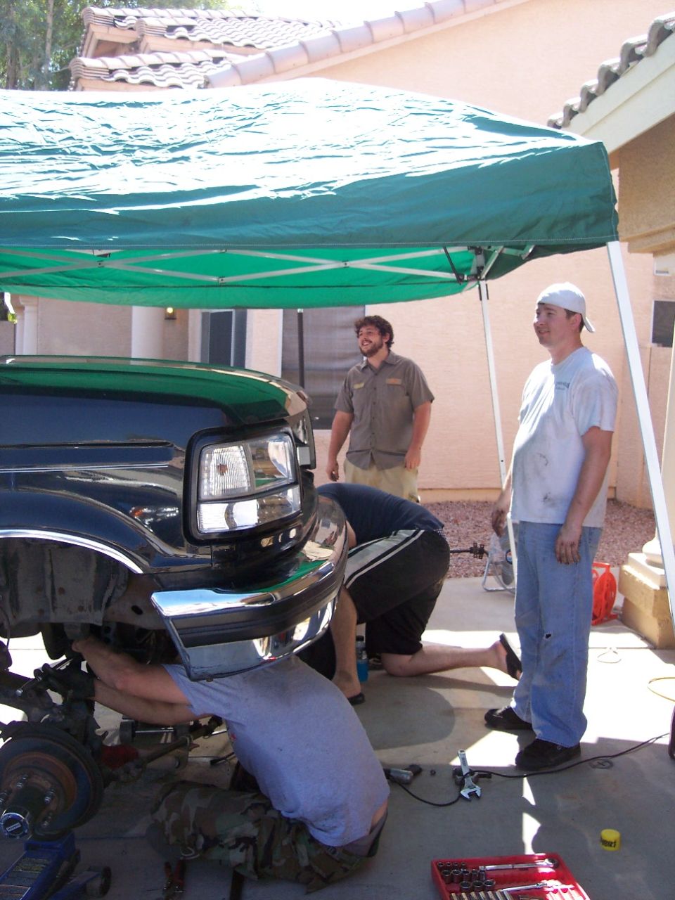 Three men working on the front of a black 1996 Ford Bronco under a green canopy.