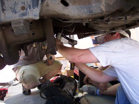 1996 Ford Bronco - Two men work underneath the chassis of a vehicle, with one man using a tool on a suspension component. The vehicle appears to be a truck or…