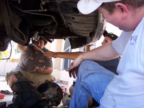 1996 Ford Bronco - Three men work on the undercarriage of a vehicle, with one man using a wrench on a suspension component. A disassembled brake rotor and hub…