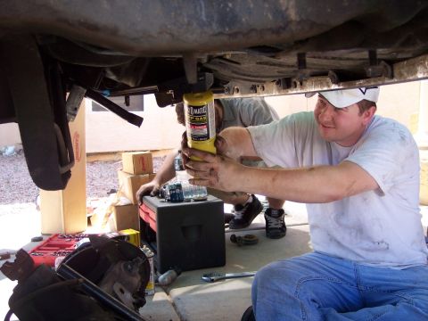 1996 Ford Bronco - A man in blue jeans and a dirty white t-shirt works underneath a vehicle, holding a yellow propane torch. Tools and parts are scattered on…