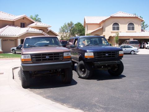 1996 Ford Bronco - Two lifted Ford Broncos are parked in front of a residential house. The Bronco on the left is brown, and the Bronco on the right is dark bl…