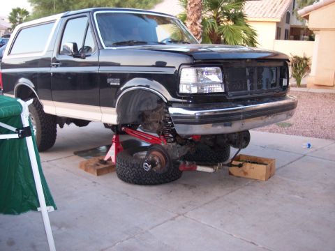 1996 Ford Bronco - A black and tan two-tone full-size Ford Bronco sits on jack stands and wood blocks in a driveway with the right front wheel removed, exposi…