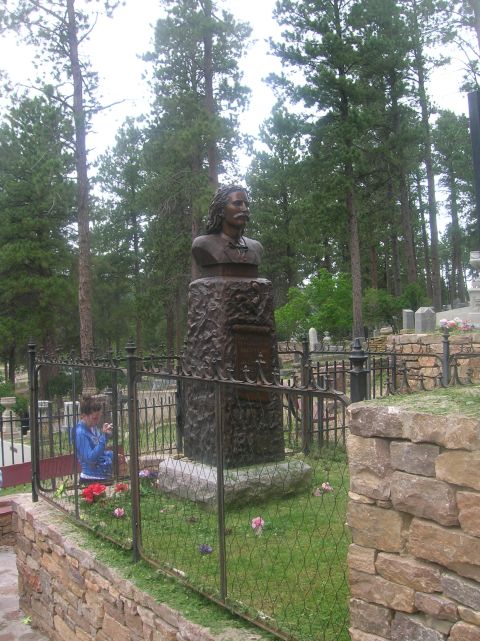 1996 Ford Bronco - A bronze bust of a man with a mustache stands on a stone pedestal in a cemetery, surrounded by pine trees. A young woman in a blue hoodie i…