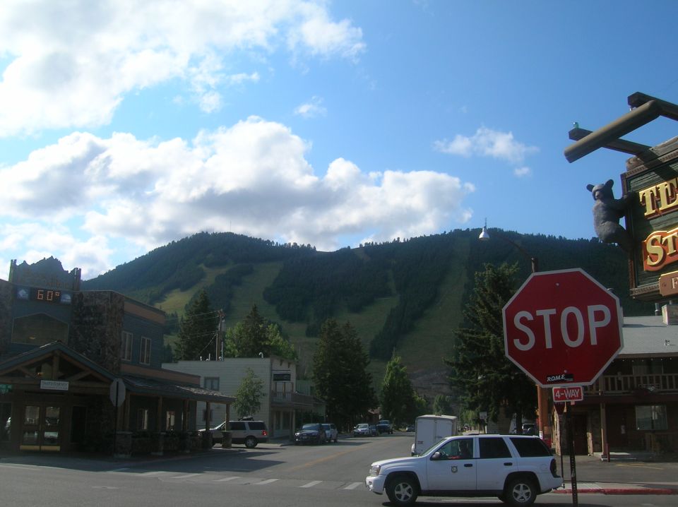 Street view in a mountain town with buildings, trees, a stop sign, and a white SUV