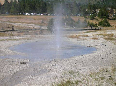 1996 Ford Bronco - A geyser sprays water into the air in a geothermal area, with a parking lot and buildings visible in the distance.