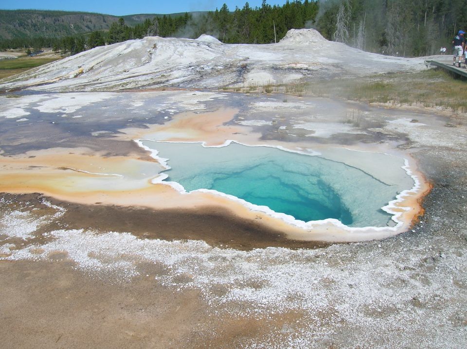 Turquoise geothermal pool with mineral deposits and geyser in background