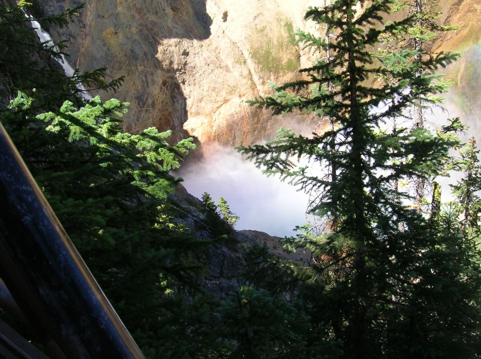 Misty waterfall in a canyon with pine trees and a wooden railing.