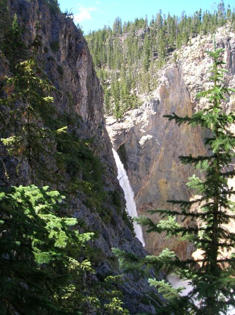 1996 Ford Bronco - A tall waterfall cascades down a rocky canyon, surrounded by dense pine forests under a bright blue sky.