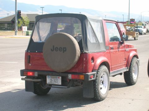 1996 Ford Bronco - The rear of a red Suzuki Samurai soft top convertible is shown in a street setting. A spare tire cover with the word "Jeep" is mounted on t…