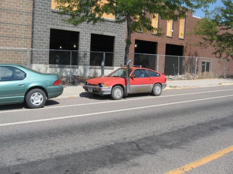 1996 Ford Bronco - A red two-door hatchback with a prominent vertical exhaust pipe is parked next to a teal sedan on a paved street in front of a brick buildi…