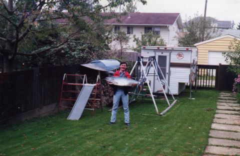 1989 Ford Bronco - A person stands in a grassy backyard holding a large fish. Behind them is a small camper trailer and a swing set.