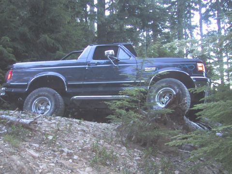 1989 Ford Bronco - A dark blue Ford Bronco navigates a rough, wooded terrain.