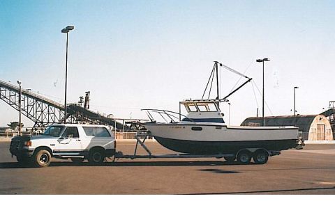 1991 Ford Bronco - A white 1991 Ford Bronco tows a white and blue boat on a trailer down a paved area with industrial structures in the background.