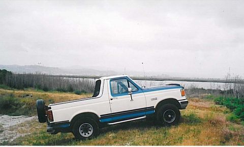 1991 Ford Bronco - A white and blue 1991 Ford Bronco is parked on a grassy hill overlooking a body of water and distant hills under a cloudy sky. The vehicle…