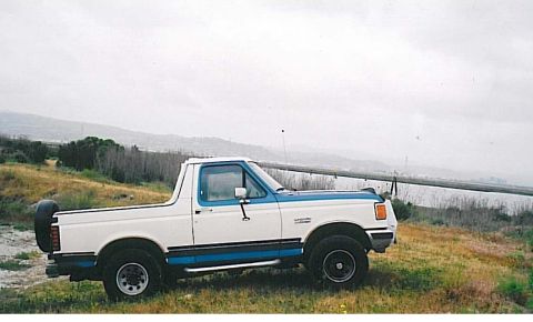 1991 Ford Bronco - A white and blue 1991 Ford Bronco is parked on a grassy hill overlooking a body of water and distant hills under an overcast sky.