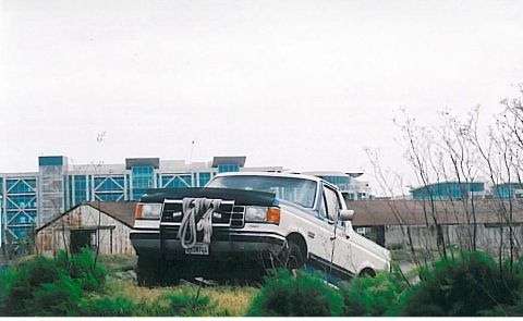 1991 Ford Bronco - A white and blue Ford pickup truck is parked on a grassy incline in front of some dilapidated buildings and modern structures in the backgr…