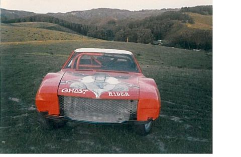 1991 Ford Bronco - A red modified race car with "GHOST RIDER" text on the hood is parked on a grassy hill with rolling hills and trees in the background.