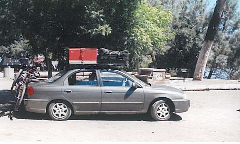 1991 Ford Bronco - A gray sedan is parked outdoors with two bicycles mounted on a rear rack and luggage on the roof rack. A red cooler is visible on top of th…