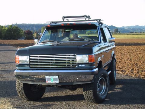 1989 Ford Bronco - This black Ford Bronco is shown from a three-quarter front angle, parked on a gravel surface with a field in the background.