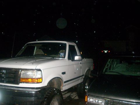 1992 Ford Bronco - A white Ford pickup truck is parked at night, possibly during rain or snow, next to a dark-colored vehicle. The front of the white truck is…