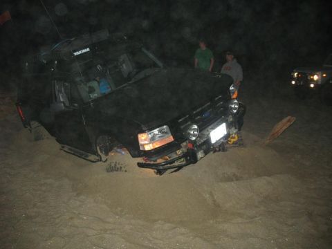 1996 Ford Bronco - A dark-colored SUV with a roof rack (Yakima branding) tilted at an angle, front end buried in sand at night, with two people standing besid…