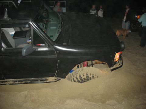 1996 Ford Bronco - A black Ford Bronco is stuck in the sand at night. The front right tire and suspension are visible, partially buried in the sand.