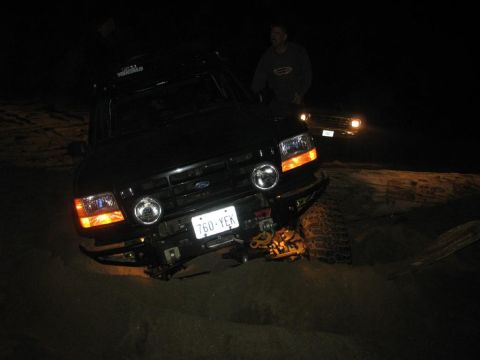 1996 Ford Bronco - A black 1996 Ford Bronco is stuck in the sand at night, with its headlights and hazard lights illuminated. Another vehicle is visible in th…