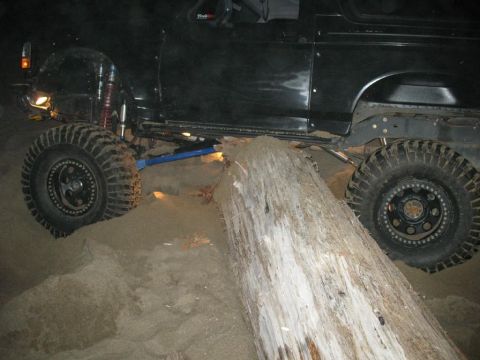 1996 Ford Bronco - A modified black 1996 Ford Bronco navigates over a large fallen log on a sandy terrain. The vehicle features large off-road tires and visib…