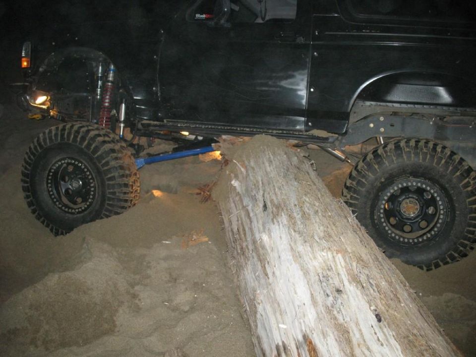 Modified black 1996 Ford Bronco driving over a large log on sand.