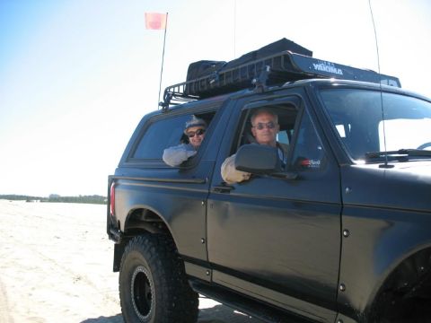 1996 Ford Bronco - Two people are in a black Ford Bronco on a sandy beach under a clear sky. A pink flag is mounted on a pole above the vehicle's roof rack.